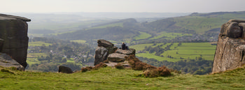 A view from Curbar Edge This landscape photograph features a panoramic view from Curbar Edge, an elevated escarpment in the Peak District, England. The image was captured in the late morning during autumn, as indicated by the subdued sunlight and the brown tones beginning to appear in the grassy foreground. The vantage point provides an extensive rural overview of the surrounding countryside in the Peak District National Park, with patchwork fields and wooded hills stretching into the distance. The village nestled below, typical of rural England, is visible with clusters of houses and winding roads. Rocky outcrops, characteristic of Curbar Edge, frame the scene, while a solitary figure sits on one of the rocks, emphasizing the scale of the landscape. This photograph highlights the natural beauty of this region in the United Kingdom, known for its dramatic viewpoints and open rural spaces.